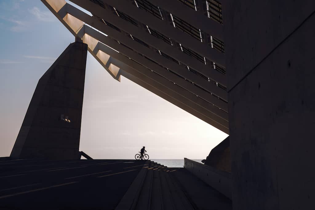 silhouette of a cyclist under a very famous steel and concrete structure of the Forum of Barcelona. The biker is next to some stairs and in the background you can see the sea, the sky is clear (silhouette of a cyclist under a very famous steel and con