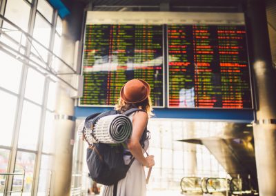 Theme travel public transport. young woman standing with back in dress and hat behind backpack and camping equipment for sleeping, insulating mat looks schedule on scoreboard airport station